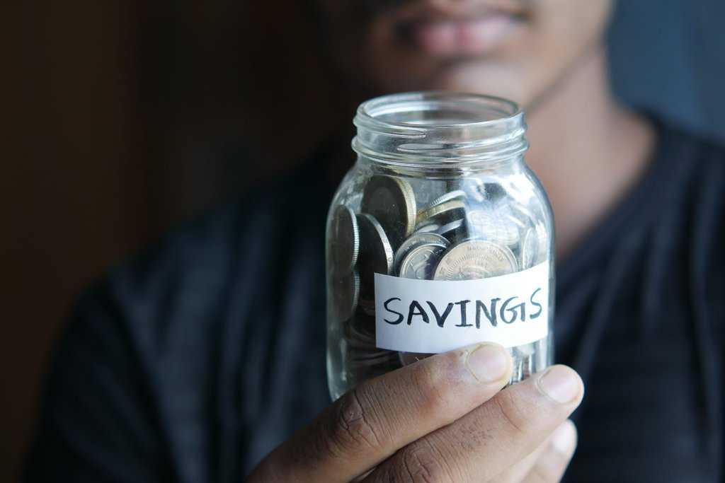 A man holding a jar of coins, displaying the word savings, while emphasizing the importance of budgeting and reducing bills to manage household expenses effectively.