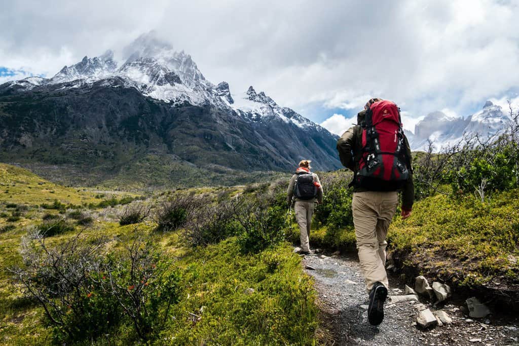 Two hikers walking along a trail with dangerous areas in the background.