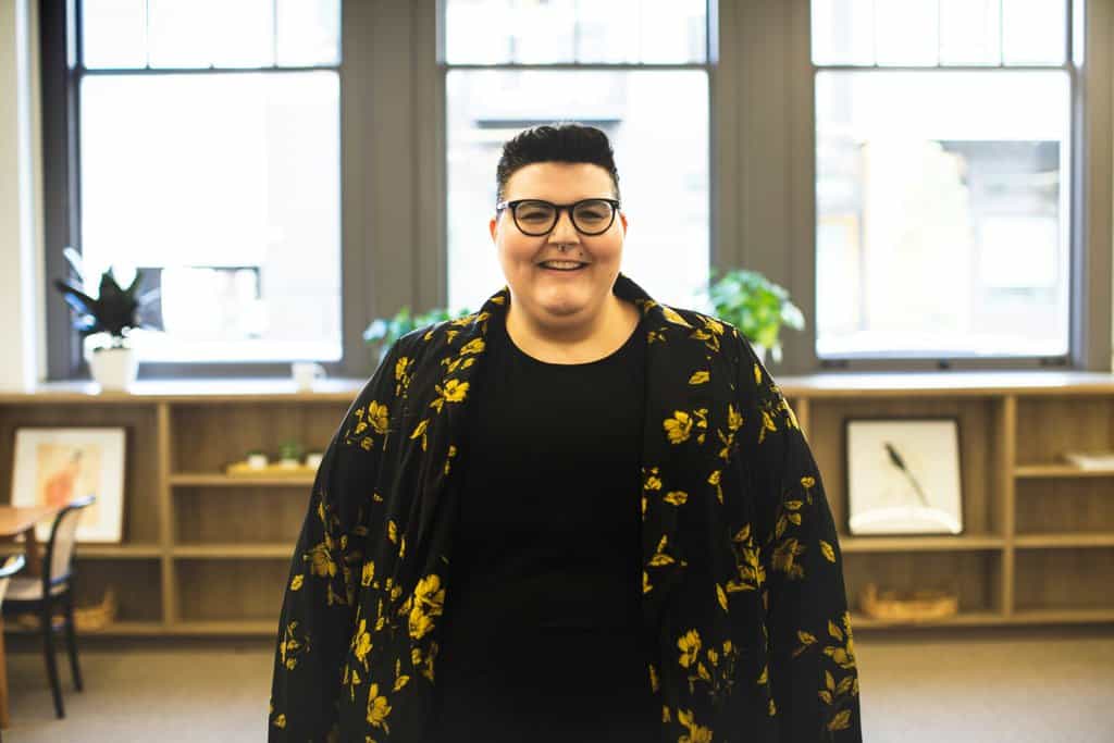 Person with short hair and glasses, wearing a patterned jacket and a Bold Over Size t-shirt, stands smiling in a well-lit room with windows and shelves in the background.