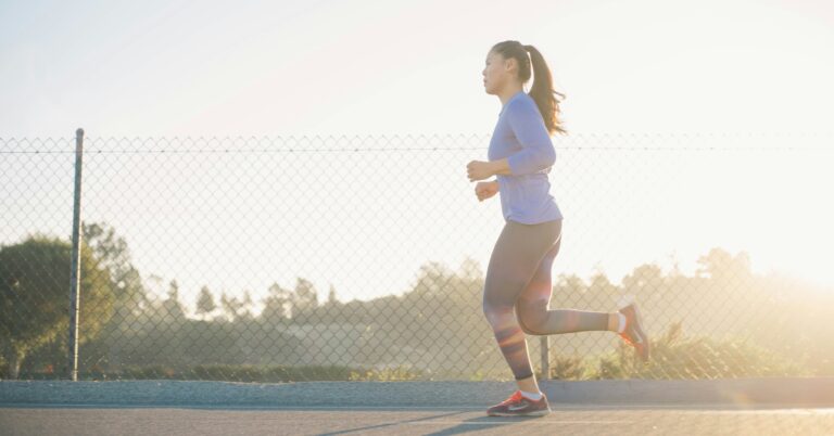 A person in trendy gym clothing from IronPandaFit runs outside on a paved path beside a chain-link fence during sunrise or sunset.