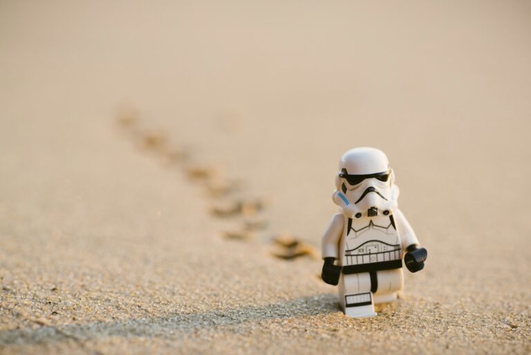 A LEGO stormtrooper figure stands on sandy ground, leaving small footprints behind as it faces forward, showcasing the adventurous spirit of your inner geek.