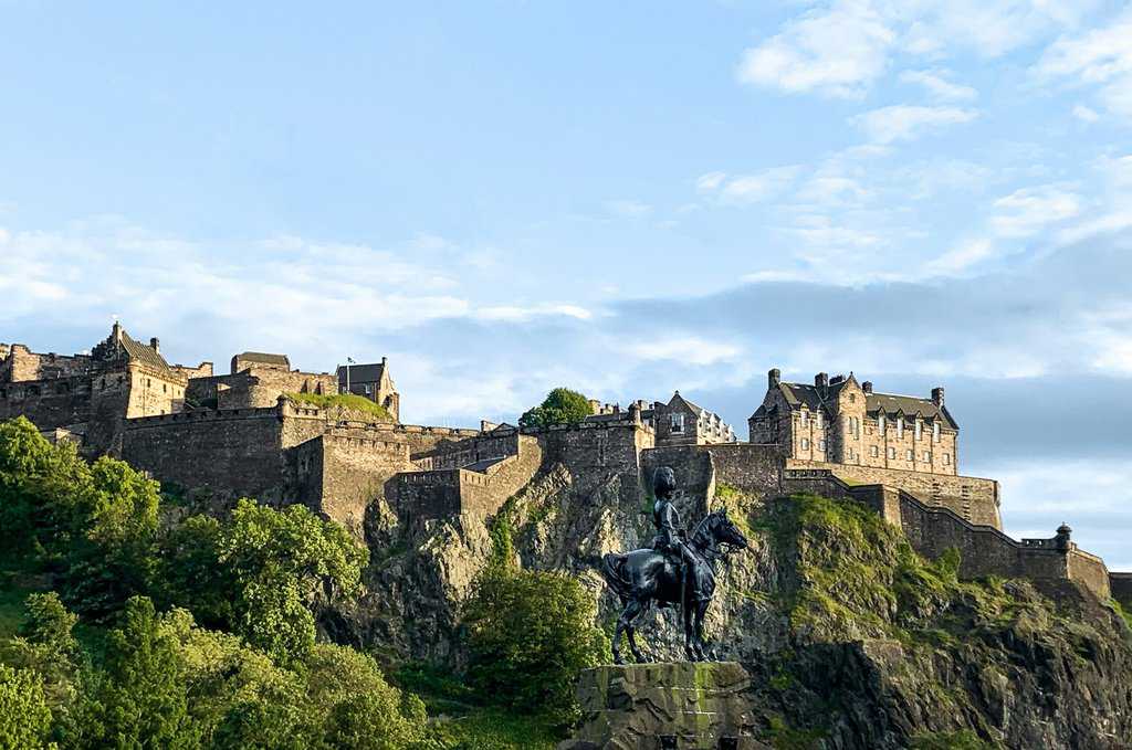 View of a castle on a hill with stone walls under a blue sky, revealing the secret charms of Edinburgh. A statue of a person on horseback stands proudly in the foreground, surrounded by lush greenery.