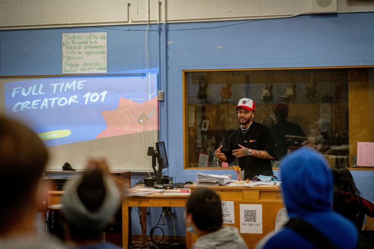 A person in stylish teacher apparel, including a hat and black shirt, is giving a presentation titled "Full Time Creator 101" to an audience in a classroom. A monitor and a desk with papers are in the background.