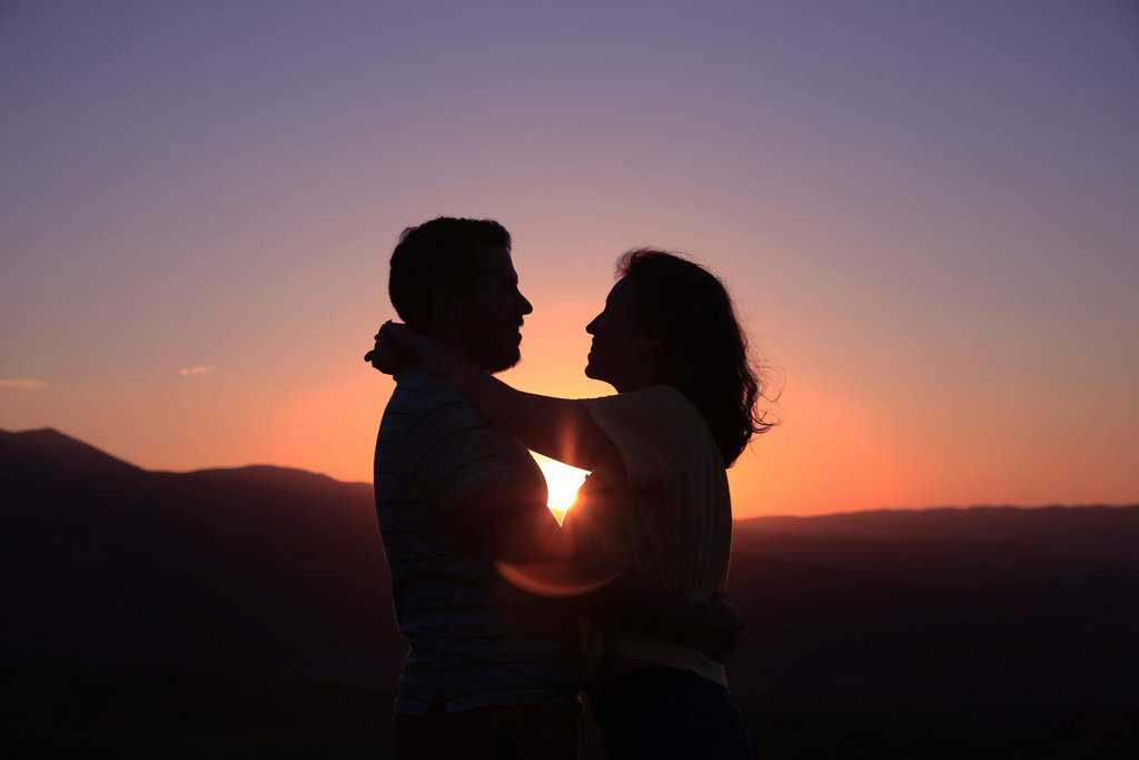A silhouette of a couple embracing at sunset, with mountains in the background and the sun partially visible near the horizon, capturing the deep connection of a long-term relationship.