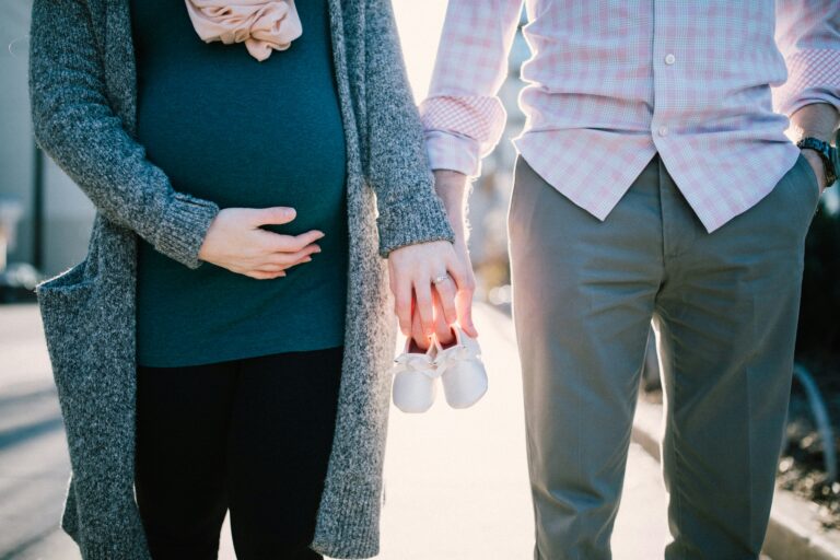 A pregnant woman and a man are standing side by side, showcasing their family pride. The woman is holding her belly, while the man holds white baby shoes. Visit Beepumpkin.com for matching clothing to celebrate moments like these.