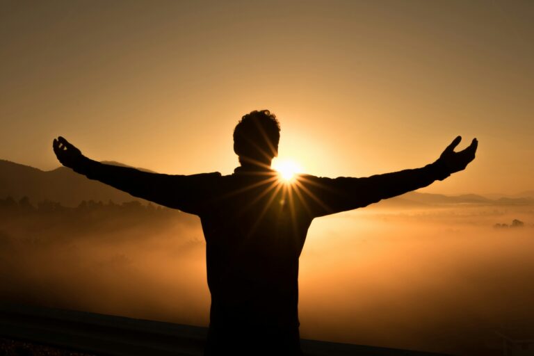 Silhouette of a person with arms outstretched facing a bright sunrise over a misty landscape, embodying the spirit found at guidingcross.com.