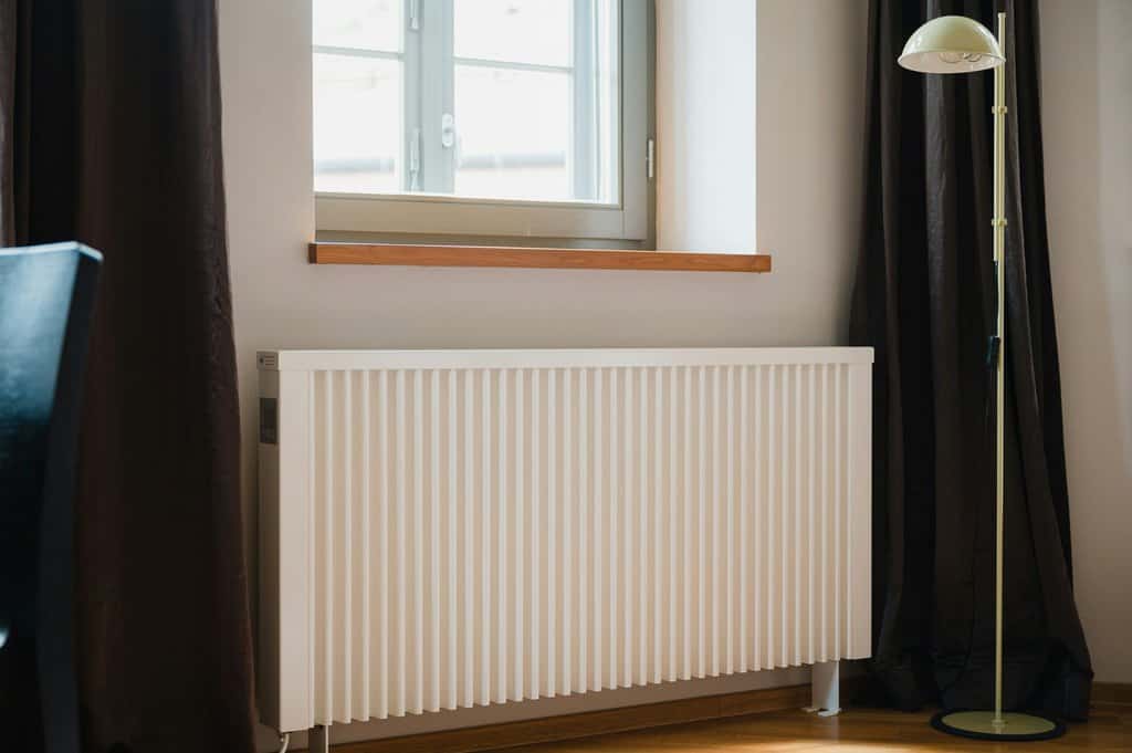 A white radiator is installed beneath a window with closed curtains on either side. To the right of the radiator stands a floor lamp, seamlessly blending into the room's auto draft design aesthetic.