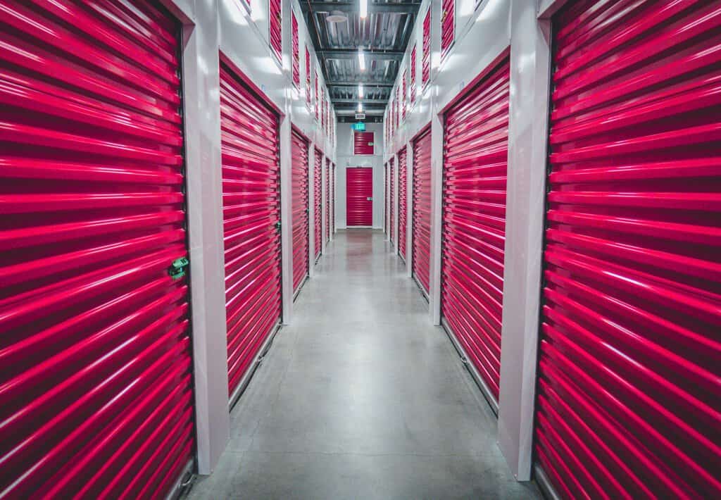 A brightly lit storage facility aisle with rows of red roll-up doors on both sides and a concrete floor, perfect for busy parents looking for summer storage tips.