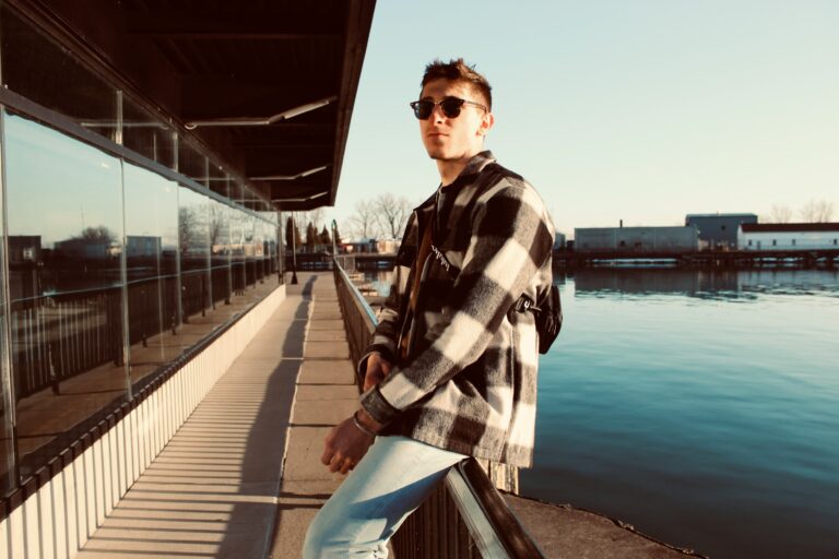 A man wearing sunglasses and a checkered jacket, exuding summer fashion vibes, leans against a railing next to a body of water on a sunny day.