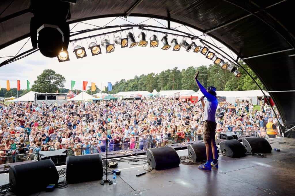 Performer on stage waving to a large outdoor festival crowd, colorful tents and flags of the Gloworm Family Festival 2024 in the background.