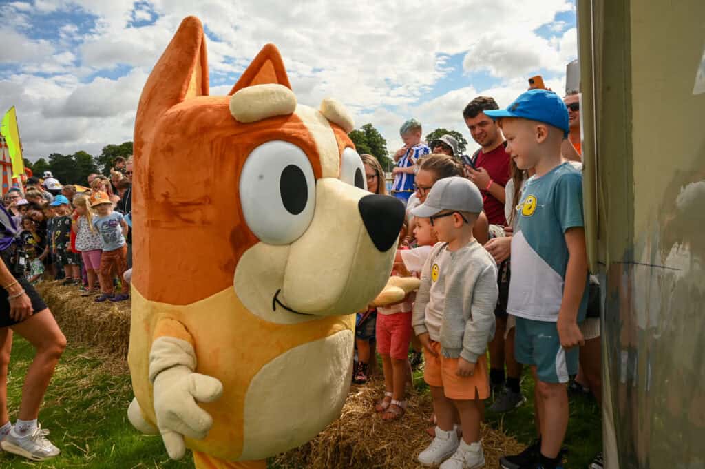 At the 2024 Gloworm Family Festival, a large, orange dog mascot interacts with children and adults at an outdoor event. The sky is partly cloudy, and there is a hay bale barrier, adding to the fun-filled activities of the day.