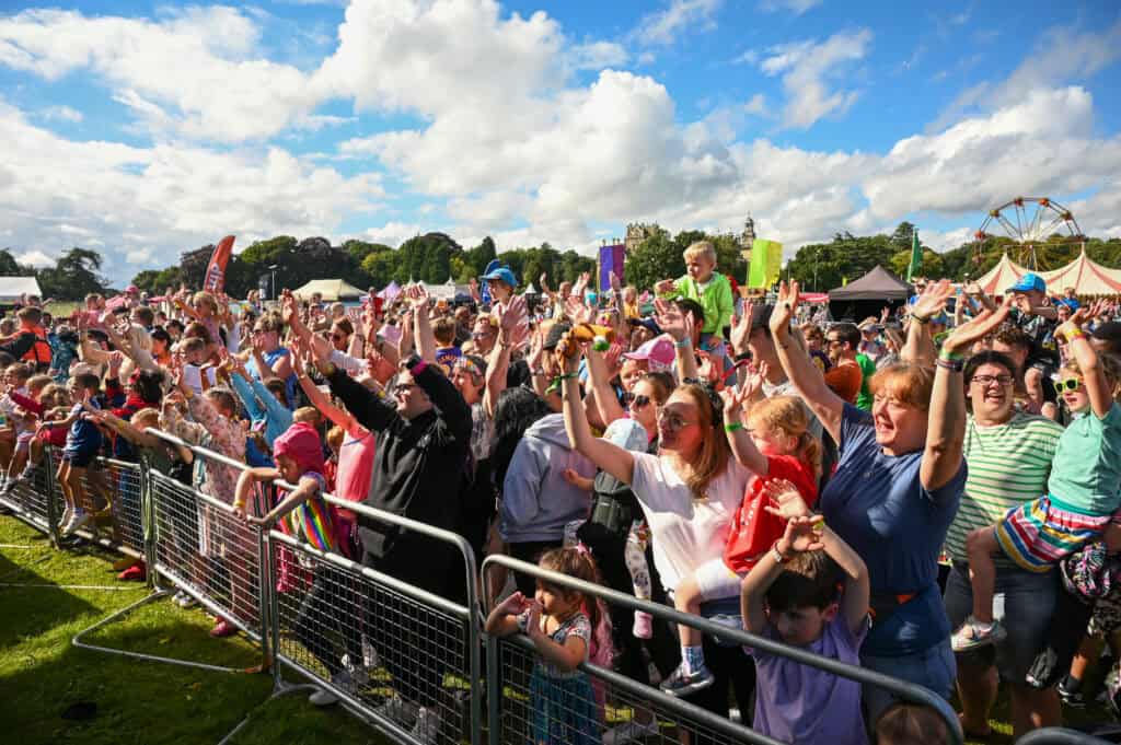 A large crowd of people, including children and adults, cheer and raise their hands behind a metal barrier at the Gloworm Family Festival 2024. On this sunny day, tents and a ferris wheel are visible in the background, promising fun-filled activities for everyone.